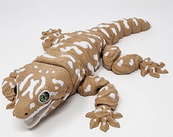 May include: A detailed, tan-colored gecko figurine with white spotted markings. The reptile has articulated limbs and a segmented body. The gecko's eyes are green, and its mouth is open. The figurine is a detailed representation of a gecko.