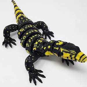 May include: A black and yellow lizard toy with a segmented body, four legs, and a long tail. The lizard has yellow spots and stripes. The toy is lying on a white surface.