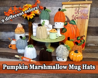 Puede incluir: Nueve gorros de taza de calabaza de ganchillo en varios colores, incluyendo verde, naranja, marrón, blanco y gris. Los gorros se muestran en un soporte de tres niveles con un rodillo y un letrero que dice "Happy Harvest".
