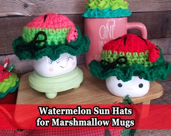 May include: Two white ceramic mugs with smiling faces are topped with crocheted watermelon sun hats. The hats are green, red, and black. The mugs are sitting on a wooden surface. "Watermelon Sun Hats for Marshmallow Mugs" is written in red text on a red background.