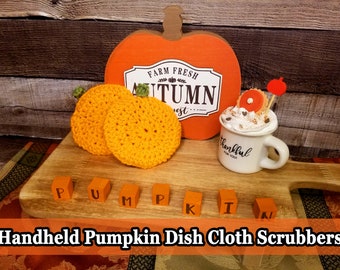 May include: Two yellow crocheted pumpkin-shaped dishcloth scrubbers sit on a wooden cutting board, next to a decorative pumpkin and a mug with whipped cream. The word "PUMPKIN" is spelled out with orange blocks.