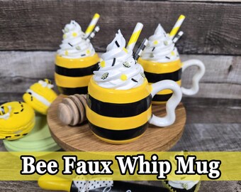 May include: Three bee-themed mugs with white whipped cream topping, yellow and black stripes, and a white handle. The mugs are decorated with yellow sprinkles and a straw. The image also includes yellow macarons and a wooden honey dipper. Text on the image reads "Bee Faux Whip Mug."