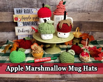 May include: A collection of crocheted apple mug hats in red and green. The hats are decorated with a brown stem and green leaf. The hats are sitting on a white mug with a happy face. The mugs are on a green pedestal. The background is a wooden wall with a sign that says "HAY RIDES Apple pick BONFIRES Raking Le CO pat"