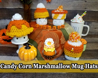 Puede incluir: Cuatro tazas blancas con gorros de maíz dulce de crochet. Las tazas tienen caras lindas y están decoradas con detalles de otoño. Las tazas están dispuestas sobre una superficie de madera con un mantel de otoño. El texto "Candy Corn Marshmallow Mug Hats" se muestra en la parte inferior de la imagen.