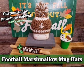 May include: A brown and white crocheted football-themed mug cozy with a white pom-pom on top. The mug cozy is sitting on a gray wooden stand. The text "TOUCHDOWN!" is on a wooden rolling pin. The background is a green and white sign that says "IT'S FALL".