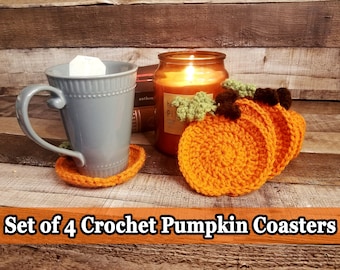 May include: Set of four crochet pumpkin coasters. The coasters are orange with green and brown accents, resembling miniature pumpkins. A blue mug with a tea bag sits on one coaster. A lit candle and books are in the background.