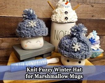 May include: A cozy, blue knit hat with a snowflake accent and a pom-pom, designed to fit a marshmallow mug. The hat is displayed on a white mug with a smiling face. A rolling pin with the text "Winter Wonderland" is also visible.