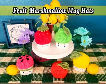 May include: Five crocheted mug hats in the shape of fruit. The hats are red, pink, green, yellow, and orange. The hats are sitting on a white tiered stand and a green and white checkered surface. The text "Fruit Marshmallow Mug Hats" is at the top of the image.