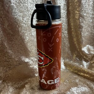 May include: A maroon water bottle with a black lid and handle. The bottle features a team logo and the word "AGE" in white. The design includes white Xs and arrows. The bottle is on a gold sequined surface.