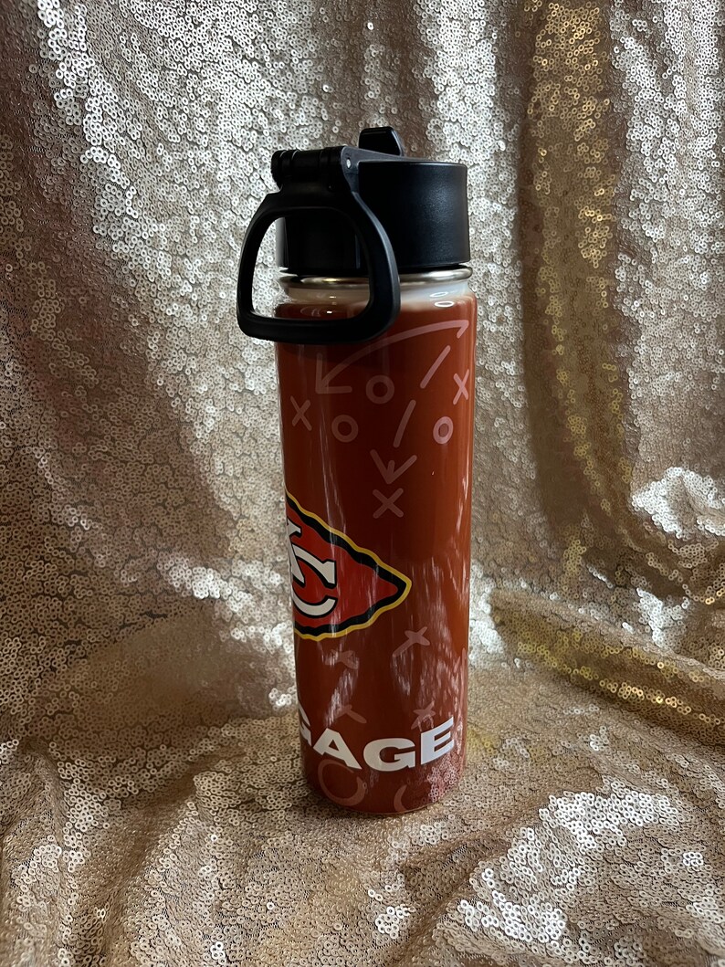 May include: A maroon water bottle with a black lid and handle. The bottle features a team logo and the word "AGE" in white. The design includes white Xs and arrows. The bottle is on a gold sequined surface.