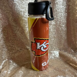 May include: A stainless steel water bottle featuring a football design with the Kansas City Chiefs logo in red and yellow. The bottle has a black lid with a handle and the word "GAME" is visible.