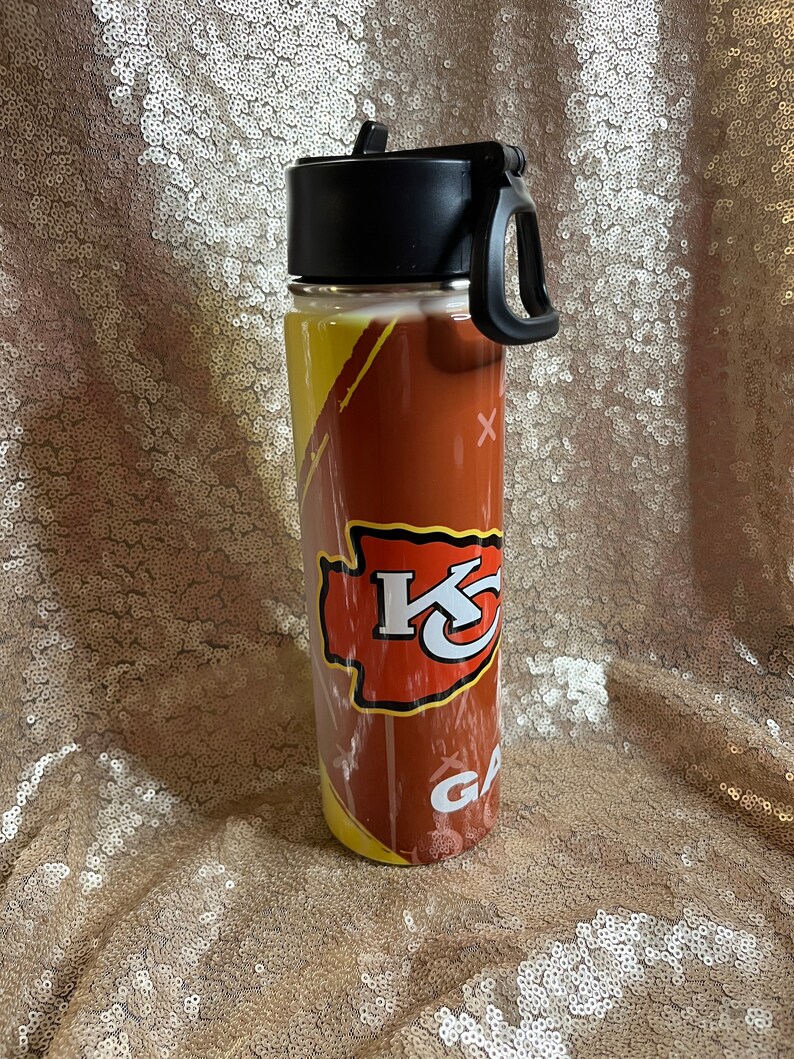 May include: A stainless steel water bottle featuring a football design with the Kansas City Chiefs logo in red and yellow. The bottle has a black lid with a handle and the word "GAME" is visible.