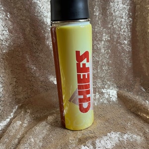May include: A yellow and brown water bottle with the word "CHIEFS" in red block letters. The bottle has a black lid and a clear body. The background is a gold sequined fabric.