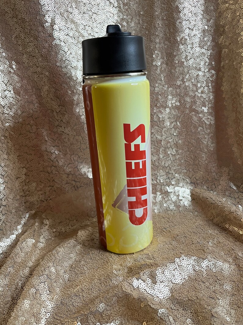 May include: A yellow and brown water bottle with the word "CHIEFS" in red block letters. The bottle has a black lid and a clear body. The background is a gold sequined fabric.