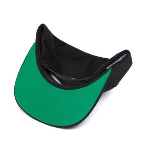 Peut inclure: Une casquette de baseball noire et verte avec une visi&egrave;re plate. La casquette est retourn&eacute;e &agrave; l'envers, montrant le dessous vert de la visi&egrave;re.