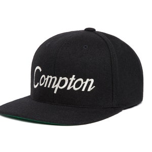 Peut inclure: Une casquette de baseball en laine noire avec un logo "Compton" brod&eacute; en blanc.