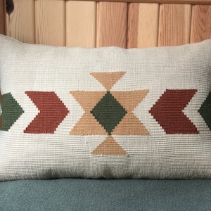 Puede incluir: Una almohada rectangular blanca con un patrón geométrico en tonos de verde, marrón y beige. El patrón presenta flechas y diamantes.