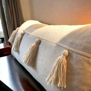 May include: A cream-colored throw pillow with a textured fabric and four white tassels. The pillow is on a bed or couch.