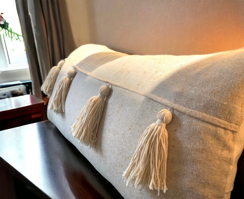 May include: A cream-colored throw pillow with a textured fabric and four white tassels. The pillow is on a bed or couch.