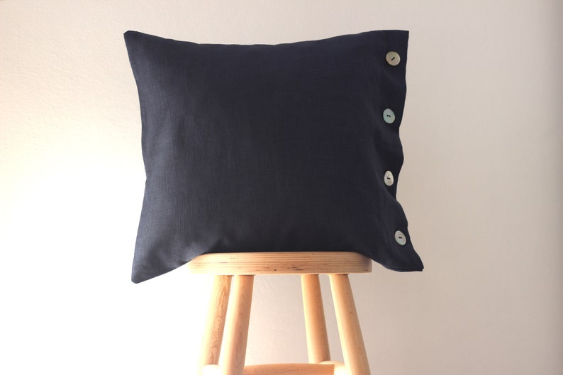 May include: A dark blue linen pillow with four white buttons down the side. The pillow is sitting on a wooden stool.