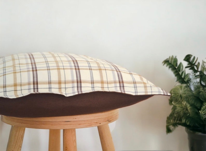 Op de afbeelding: Een bruin en beige geruit kussen met een bruine onderkant. Het kussen ligt op een houten kruk.