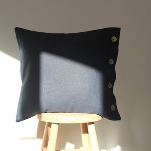May include: A dark blue linen pillow with four light brown buttons on the side. The pillow is sitting on a wooden stool.