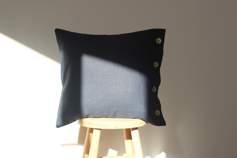 May include: A dark blue linen pillow with four light brown buttons on the side. The pillow is sitting on a wooden stool.
