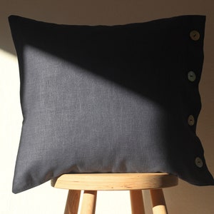 May include: A dark gray linen pillow with four wooden buttons down the side. The pillow is sitting on a wooden stool.