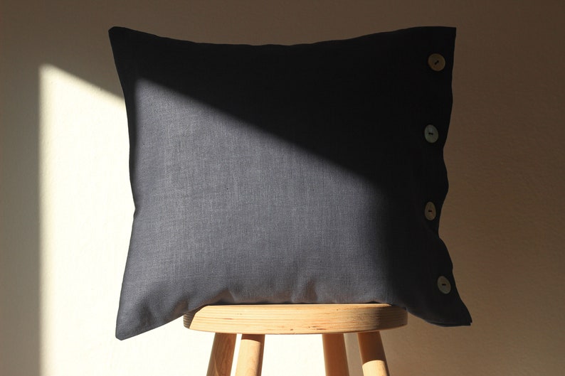 May include: A dark gray linen pillow with four wooden buttons down the side. The pillow is sitting on a wooden stool.