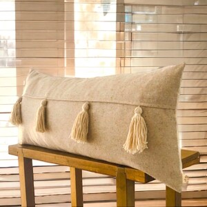 May include: A long, rectangular beige pillow with four white tassels. The pillow is sitting on a wooden bench.