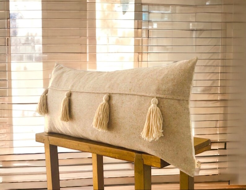 May include: A long, rectangular beige pillow with four white tassels. The pillow is sitting on a wooden bench.