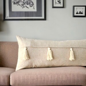 May include: A long, beige throw pillow with three white tassels. The pillow is on a light brown couch.