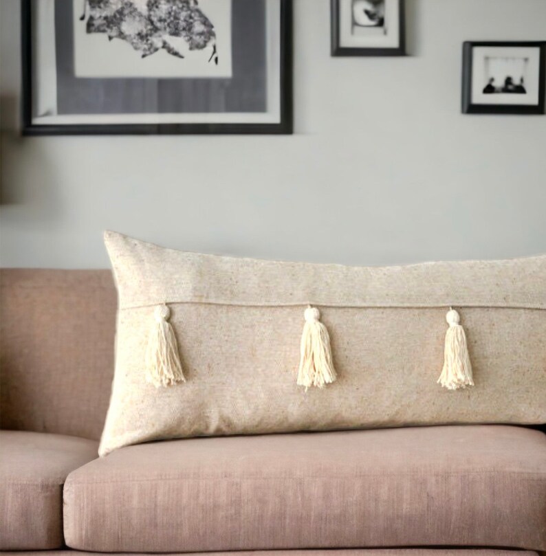 May include: A long, beige throw pillow with three white tassels. The pillow is on a light brown couch.