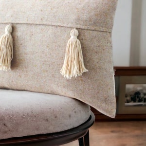 May include: A beige throw pillow with two white tassels on the front. The pillow is sitting on a light gray chair.