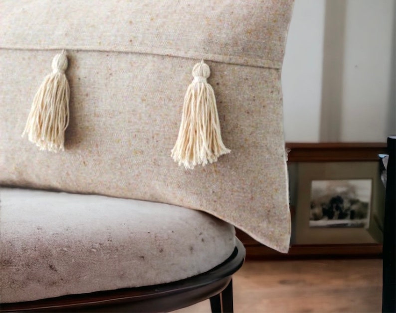 May include: A beige throw pillow with two white tassels on the front. The pillow is sitting on a light gray chair.