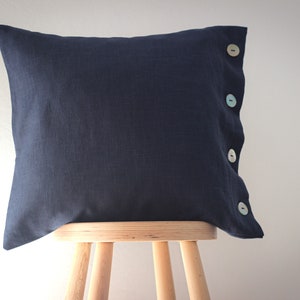 May include: A dark blue linen pillow with four pastel colored buttons down the side. The pillow is sitting on a wooden stool.