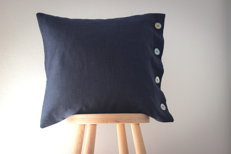 May include: A dark blue linen pillow with four pastel colored buttons down the side. The pillow is sitting on a wooden stool.