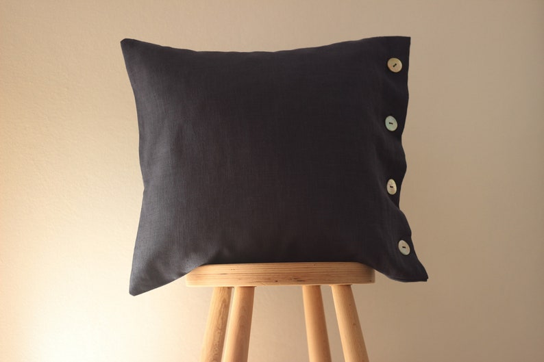 May include: A dark blue pillow with four light colored buttons down the side. The pillow is sitting on a wooden stool.