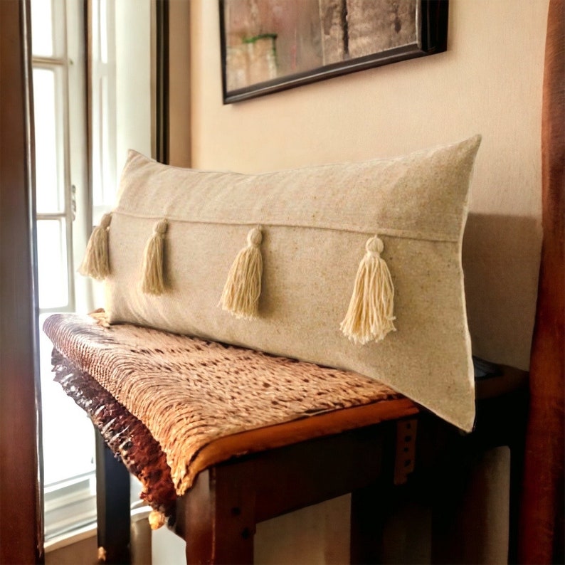 May include: A long, beige lumbar pillow with four white tassels. The pillow is sitting on a brown woven bench.