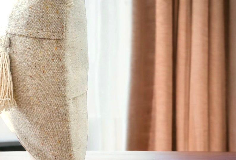 May include: A beige and white throw pillow with a tassel detail. The pillow is made of a textured fabric and has a subtle speckled pattern.