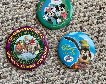 May include: Three round Disney-themed buttons. The first button is green with a white house and Mickey and Minnie Mouse in front of it. The text on the button reads "Disney Vacation Club" and "Disney's Beach Club Villas". The second button is purple with a white background and features a variety of animals including a tiger, an elephant, a monkey, a manatee, and a fish. The text on the button reads "Conservation Hero", "Disney Wildlife", "Conservation Fund", and "Disney's Animal Kingdom". The third button is blue with a white background and features Goofy in a red and yellow outfit. The text on the button reads "Heard the best kept Disney Secret?" and "Vacation Club".