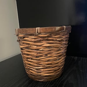 May include: A woven wicker basket with a brown and tan color scheme. The basket is cylindrical in shape and has a wide opening.
