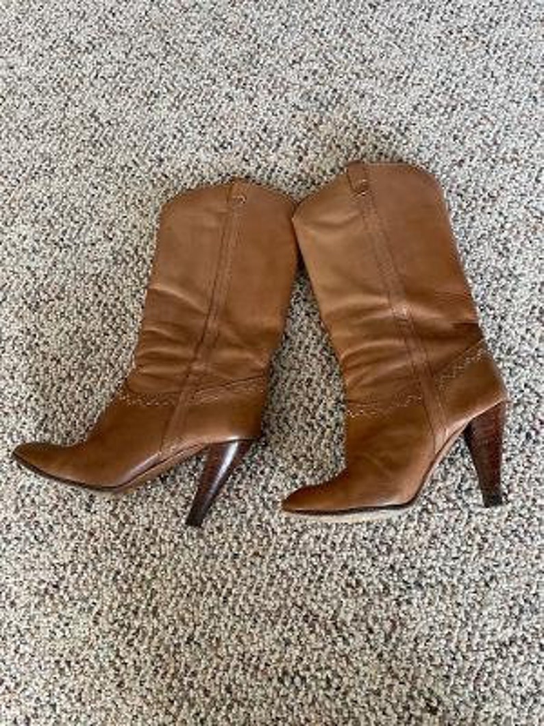 Puede incluir: Un par de botas de cuero marr&oacute;n de ca&ntilde;a alta con punta puntiaguda y tac&oacute;n de bloque.