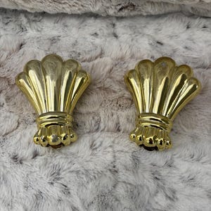 May include: Two gold-colored curtain tiebacks with a shell-like design. The tiebacks have a fan-shaped top and a decorative base. They are placed on a soft, textured, light-colored surface.