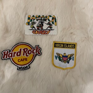 May include: Three embroidered patches on a white furry surface. The patches are for Jamaica, Hard Rock Cafe Miami, and Virgin Islands. The Jamaica patch has a green, yellow, and red color scheme with the text "Jamaica Crazy". The Hard Rock Cafe Miami patch is yellow and red with the text "Hard Rock Cafe Miami". The Virgin Islands patch is yellow and blue with the text "Virgin Islands" and a depiction of the Virgin Islands flag.