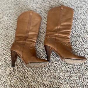 Puede incluir: Un par de botas vaqueras de cuero marr&oacute;n con punta puntiaguda y tacones altos.