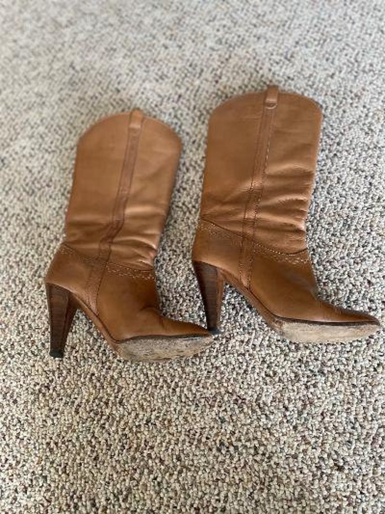 Puede incluir: Un par de botas vaqueras de cuero marr&oacute;n con punta puntiaguda y tacones altos.
