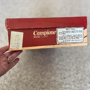 Puede incluir: Una caja de zapatos roja con las palabras "Campione Shoes" impresas en la parte delantera. La caja tiene una etiqueta blanca con informaci&oacute;n sobre los zapatos.