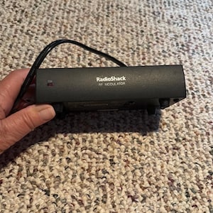 May include: A black RadioShack RF Modulator with the word "ON" and a small red light. The device has a black power cord attached. The device is on a patterned surface.
