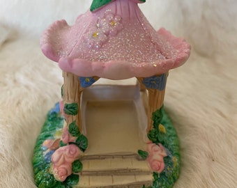 May include: A pink and green fairy garden gazebo with a ladybug on top. The gazebo has a white base with steps and is surrounded by green grass and pink flowers.